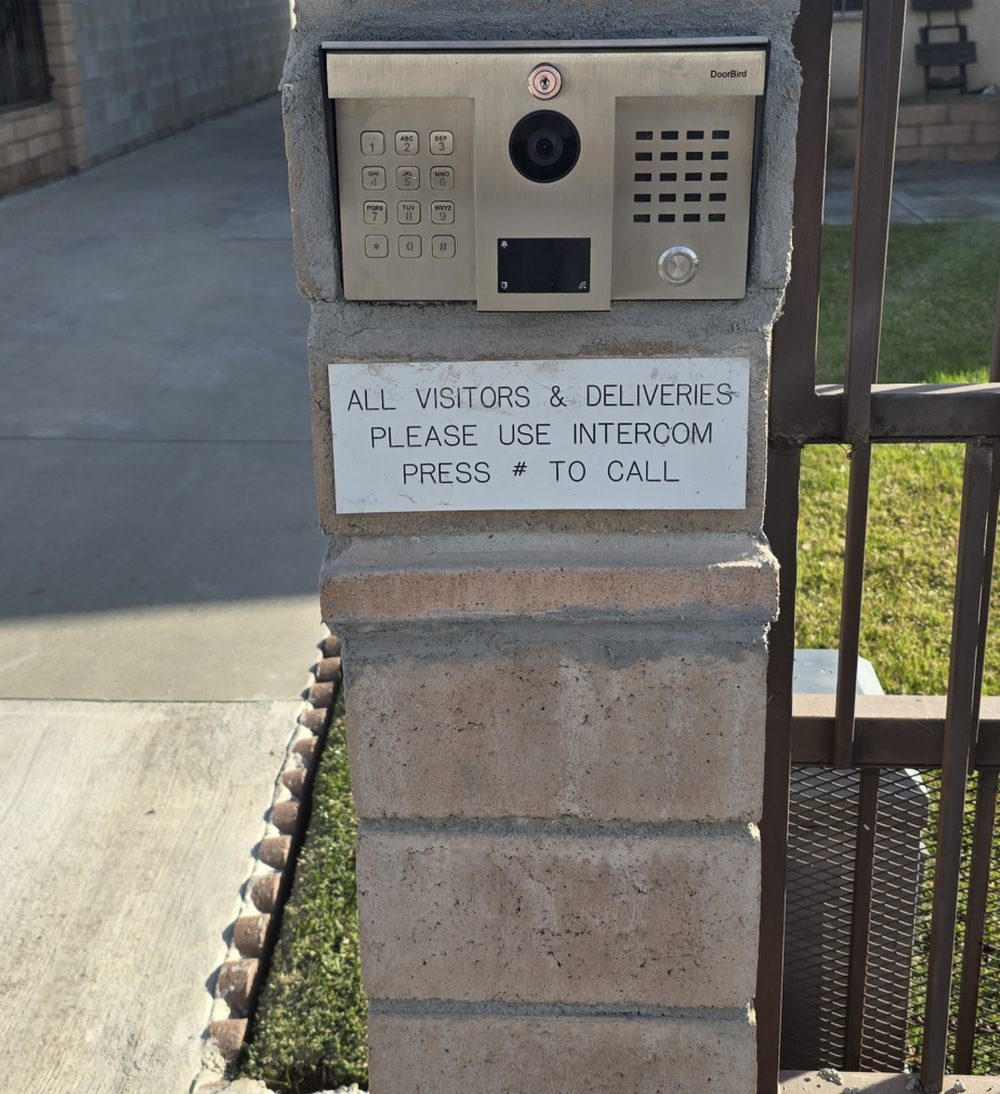 DoorBird video intercom call box with keypad, camera and RFID reader set into a block gate pillar, with a sign asking visitors and deliveries to use the intercom