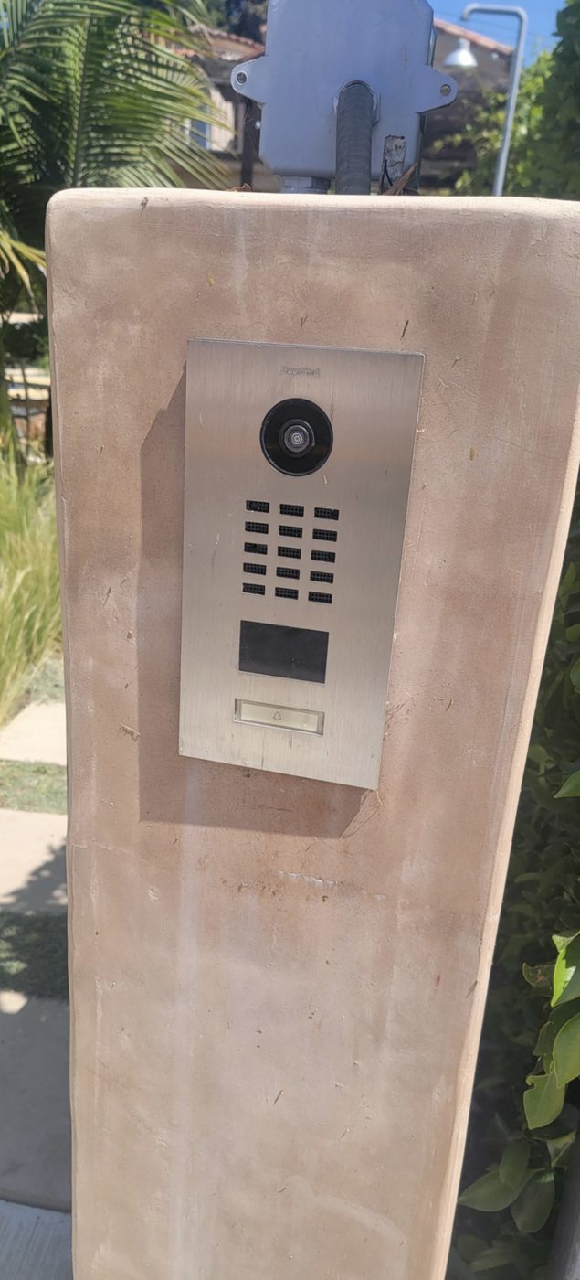 Stainless steel DoorBird IP video door station with camera, RFID reader and call button mounted on a stucco gate pillar